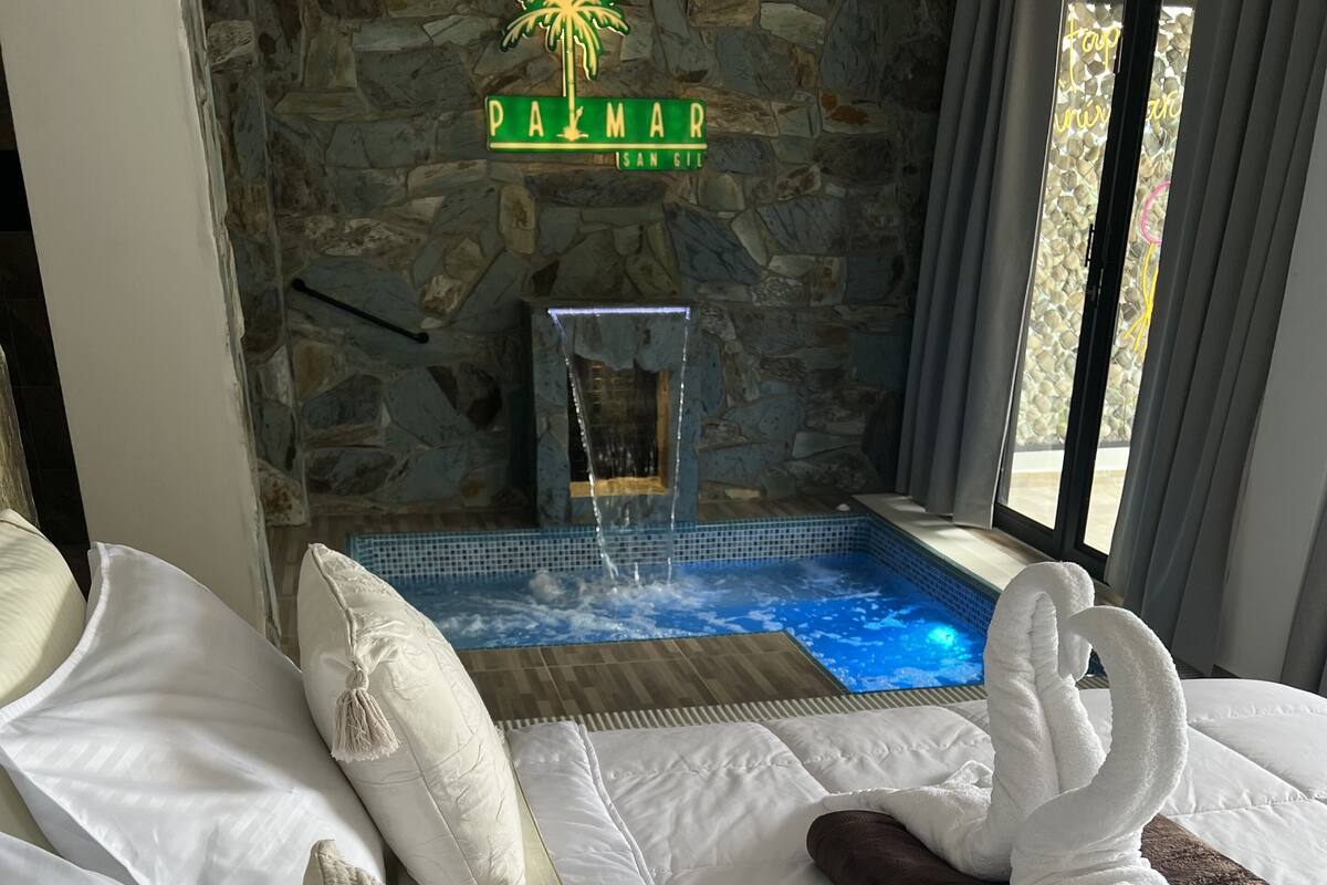 A hotel room with a stone wall, a sign reading 'PALMAR ISAN GIL,' a pool with water features, and a bed with white bedding and a macrame throw.