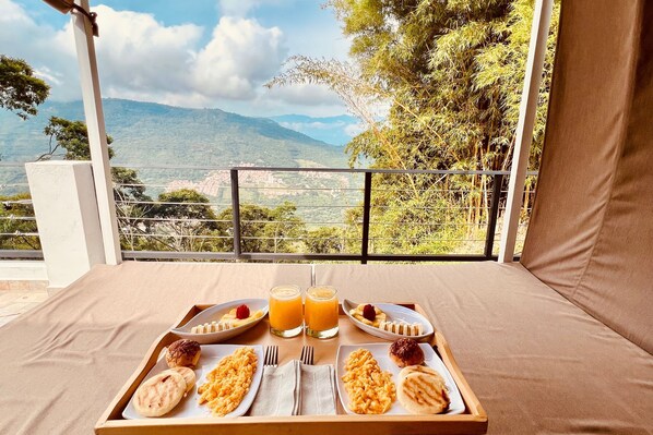 Bandeja de desayuno con panqueques, fruta y jugo de naranja en un balcón con vistas a un paisaje montañoso.