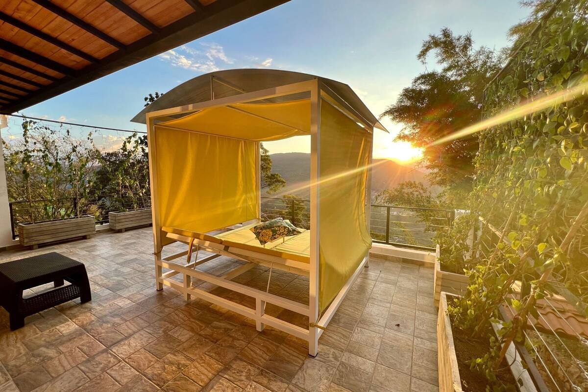 A covered outdoor seating area with a bench and a small stool, surrounded by greenery and a view of the landscape.