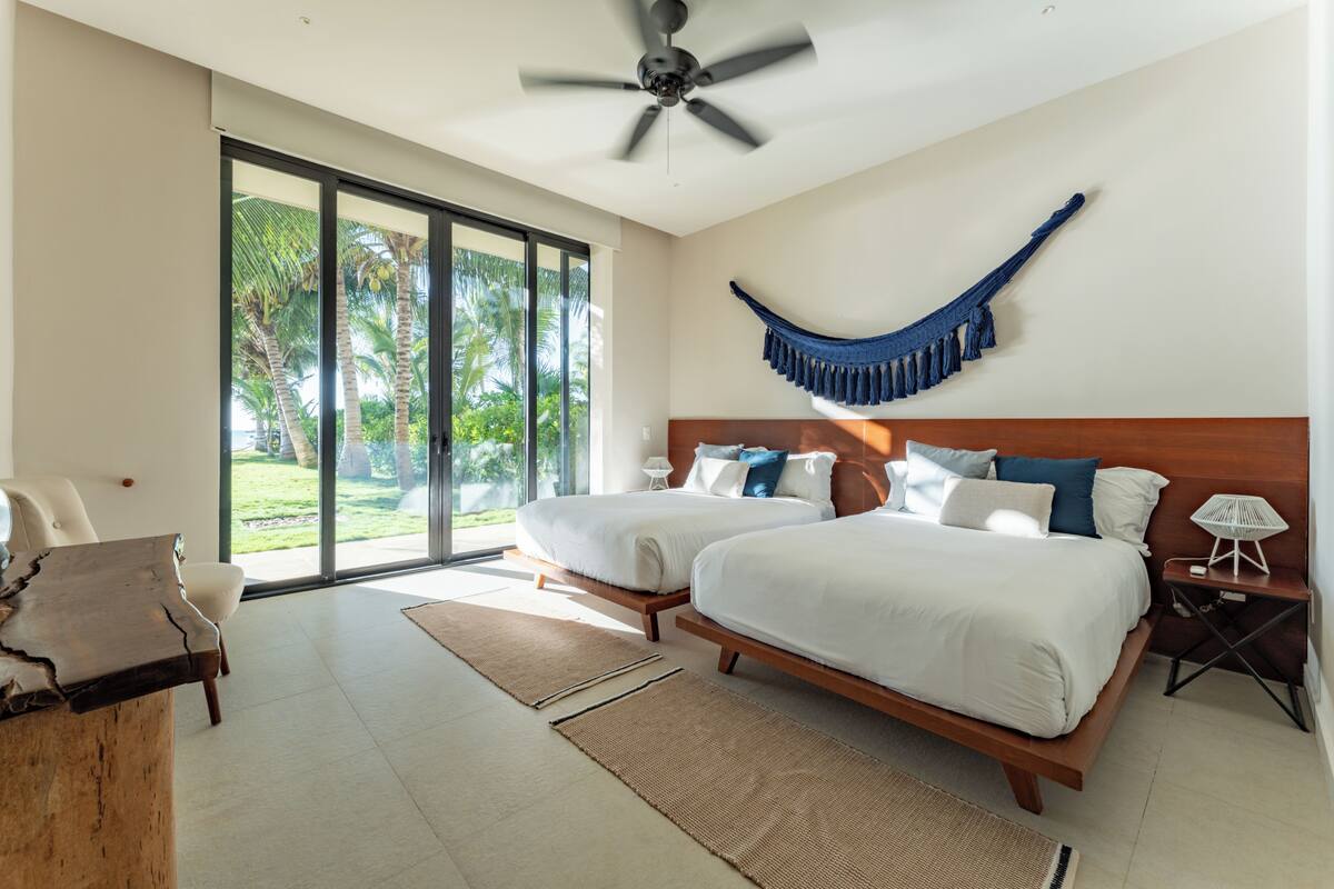 A bedroom with two beds, a ceiling fan, a hammock, and a view of palm trees.
