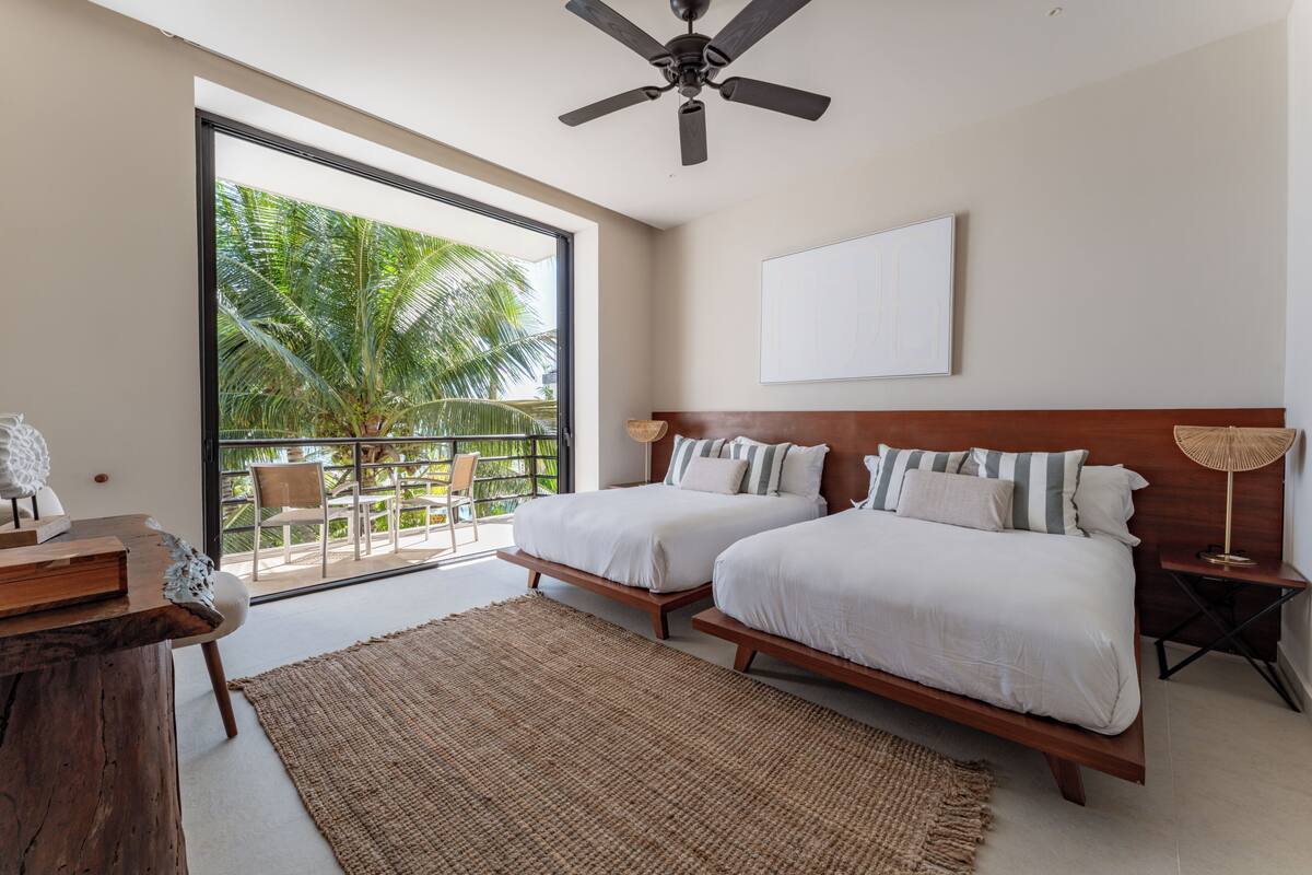 A bedroom with two beds, a ceiling fan, a large window with a view of palm trees, and a balcony with chairs.
