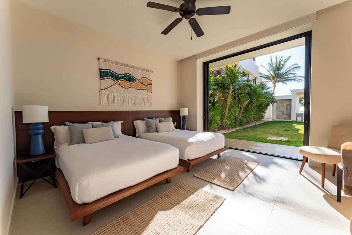A bedroom with two beds, a ceiling fan, a large window, and a view of a garden.
