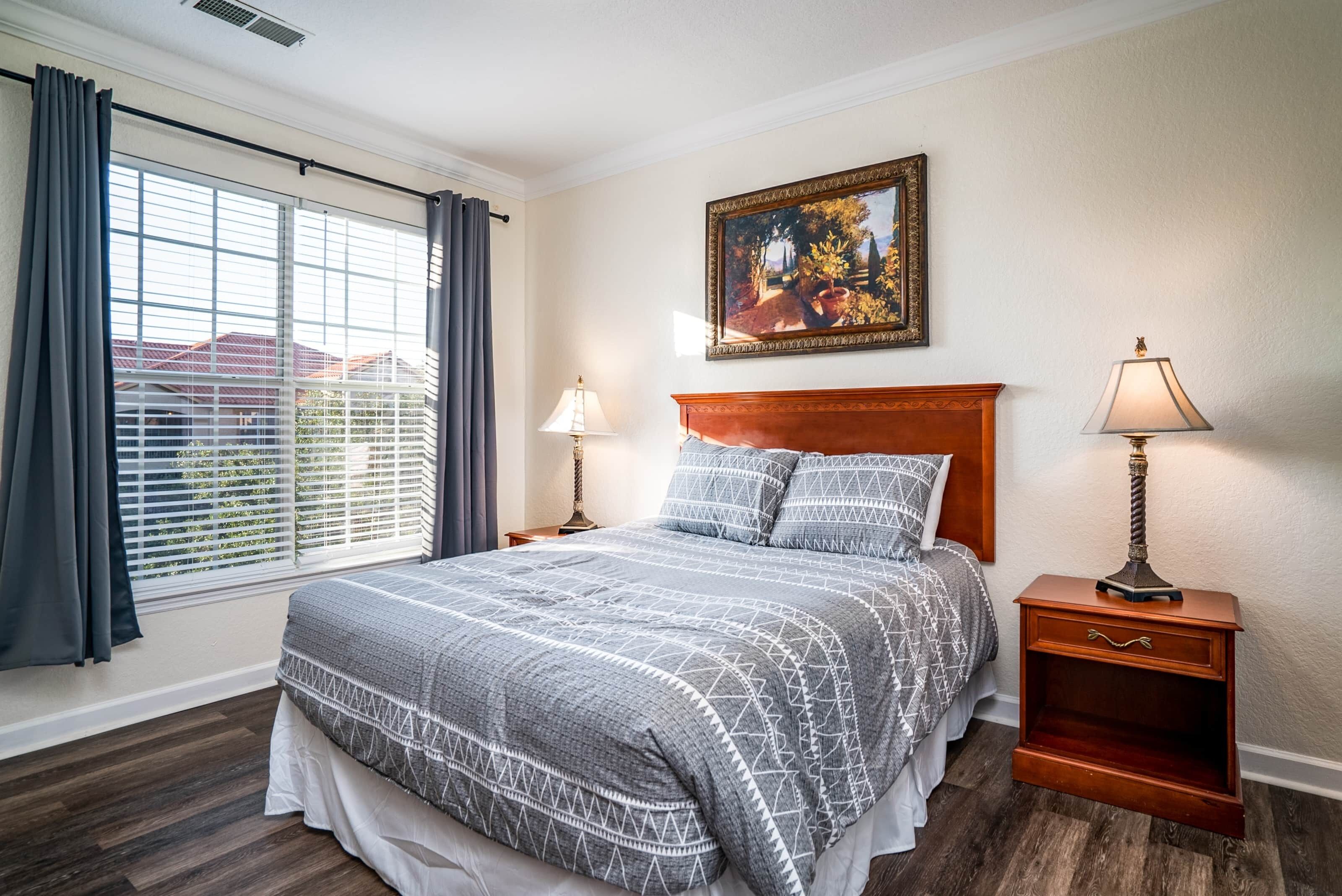 A bedroom with a large bed, two bedside tables, a nightstand, a window with curtains, and a painting on the wall.