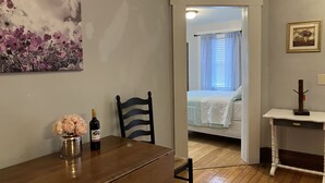 A room with a wooden table, a chair, a bed with white bedding, a painting on the wall, and a framed picture. - 2-Bedroom Apt Near Hampton Beach (Hampton)