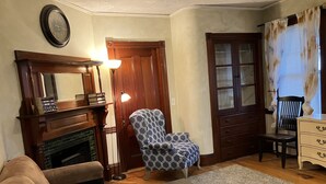 A living room with a fireplace, a patterned armchair, a wooden dresser, and a clock on the wall. - 2-Bedroom Apt Near Hampton Beach (Hampton)