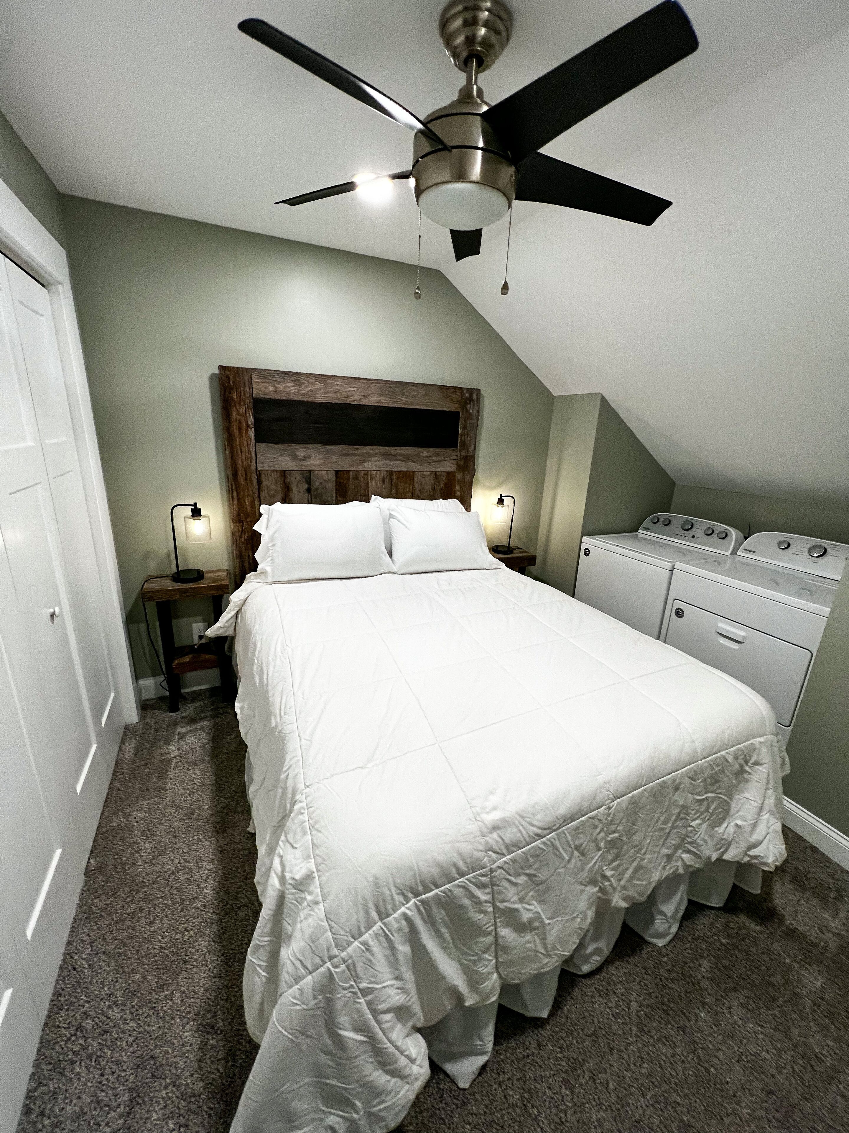 Un dormitorio con cama, ventilador de techo, mesita de noche con lámpara y lavadora.