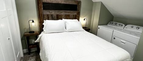 Un dormitorio con cama, ventilador de techo, mesita de noche con lámpara y lavadora.