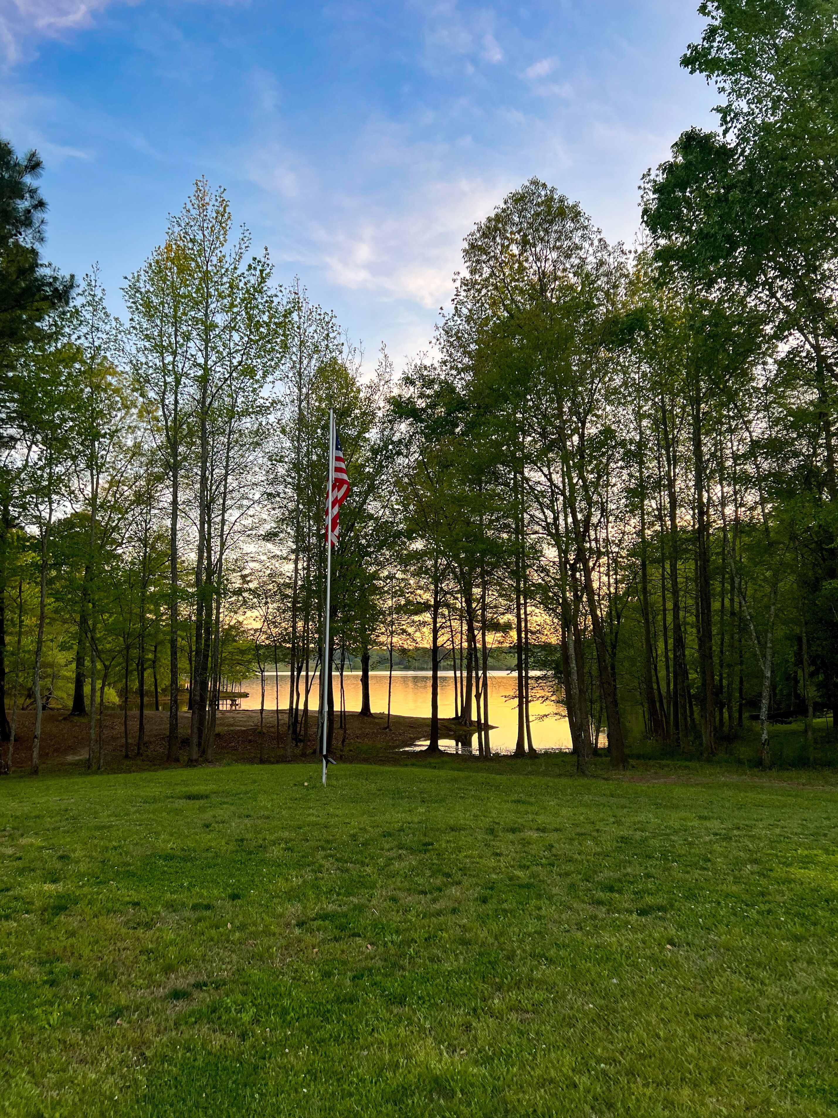 A grassy field with a flagpole and a bench near a body of water, surrounded by trees.