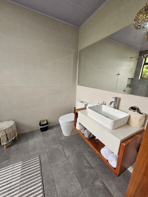 Une salle de bain moderne avec un grand miroir, deux lavabos, des toilettes et une petite table ronde recouverte d’une housse en macramé.
