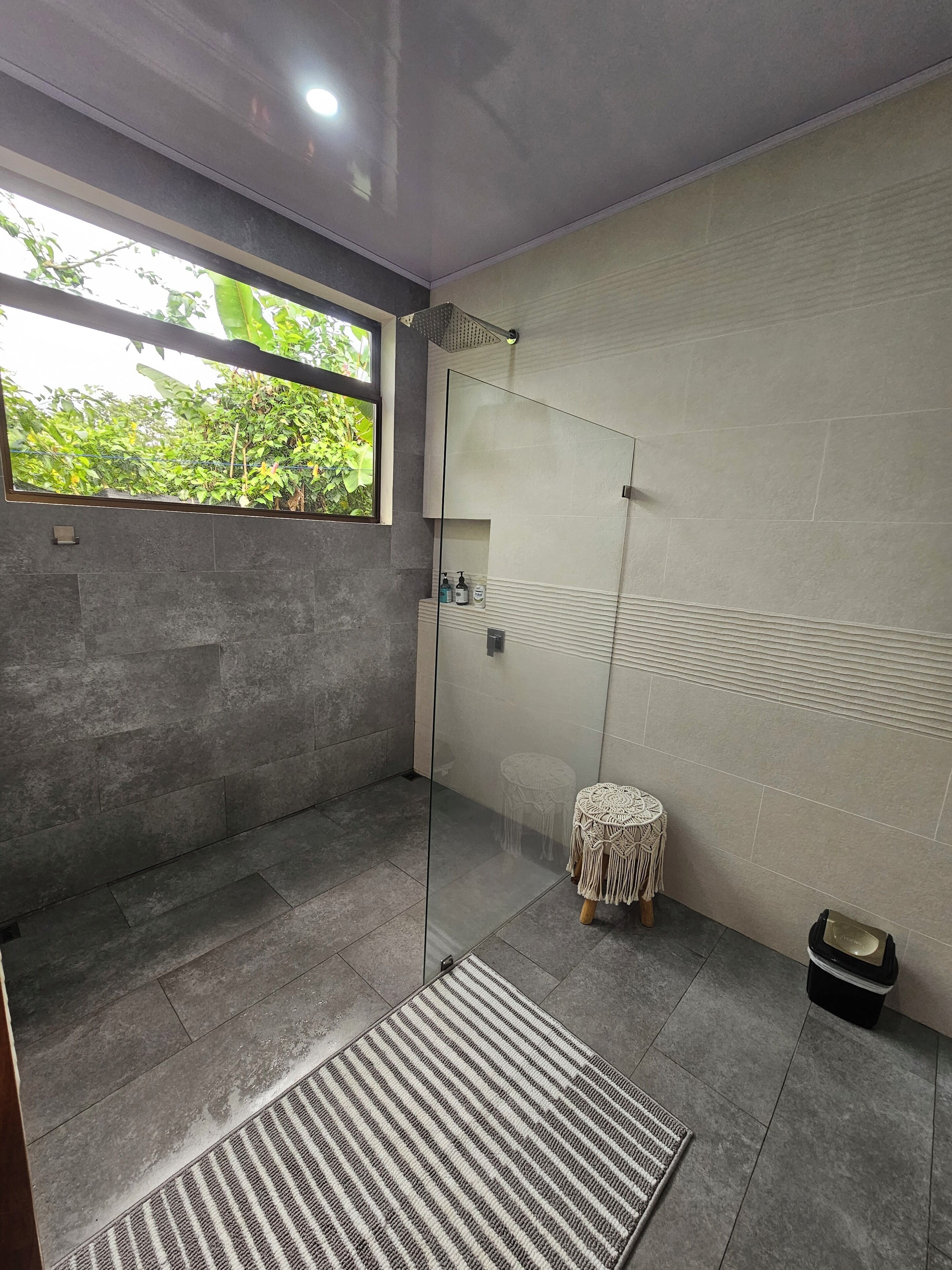 A modern bathroom with a glass shower enclosure, a small round stool, and a window with a view of greenery.