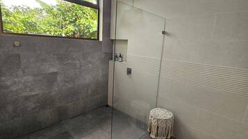 A modern bathroom with a glass shower enclosure, a small round stool, and a window with a view of greenery.