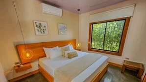 Une chambre dotée d’un grand lit, d’une tête de lit en bois, de tables de chevet, d’une commode, d’une fenêtre donnant sur un espace verdoyant et d’un climatiseur fixé au mur.