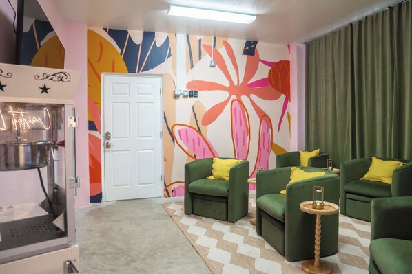 Una habitación con sofás verdes, cojines amarillos, un tapete estampado y un mural abstracto lleno de color.