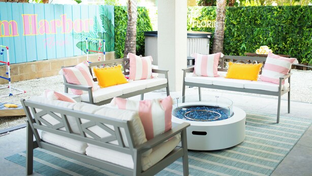 Un patio con dos sofás blancos con cojines rosa y amarillos, una mesa redonda con un brasero azul y una alfombra a rayas.