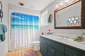Une salle de bain avec un rideau de douche représentant une scène de plage, des toilettes blanches, un lavabo avec un miroir et un meuble vert.