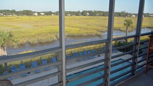 A view from a balcony overlooking a swimming pool with lounge chairs, a palm tree, and a marshy landscape with houses in the distance. - Marshside Inn 201 - Second Row (Marshfront) in Garden City Beach (Garden City)