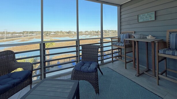 A balcony with wicker furniture, a ceiling fan, and a view of a water body and distant buildings. - Marshside Inn 201 - Second Row (Marshfront) in Garden City Beach (Garden City)