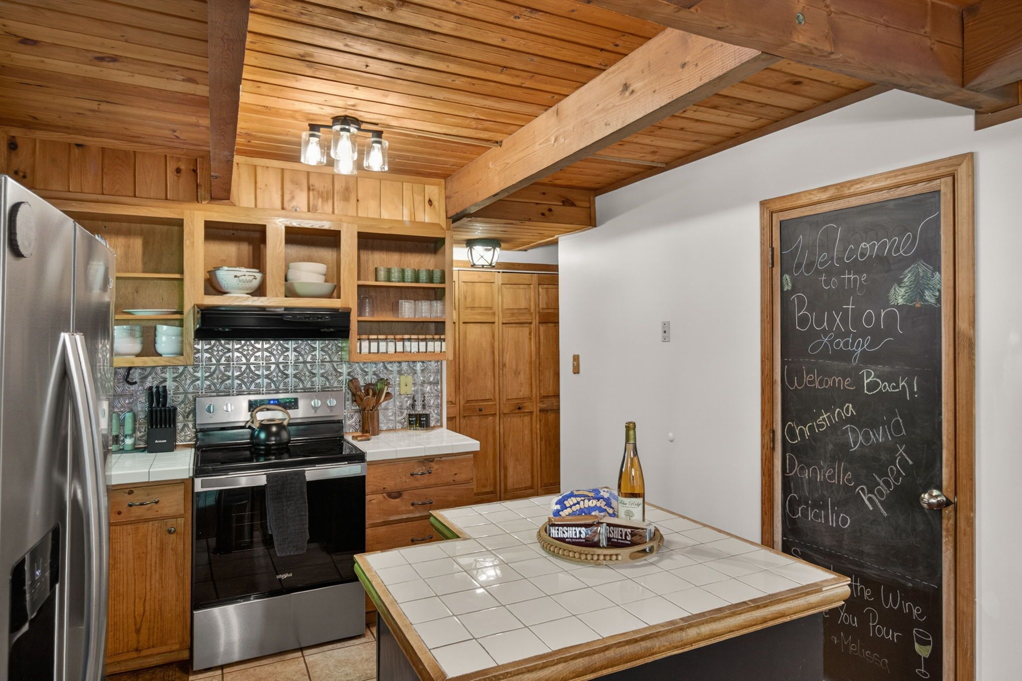 Una cocina con armarios de madera, un refrigerador de acero inoxidable, una estufa y una pizarra con nombres.
