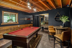 A pool table in a room with wooden beams, a bar area, and a window with a plant. - Pocono cabin where the host thought of everything, so you don’t have to! (Bushkill)