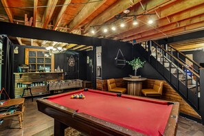 A pool table with a red felt surface, a foosball table, and a bar area with stools and bottles. - Pocono cabin where the host thought of everything, so you don’t have to! (Bushkill)