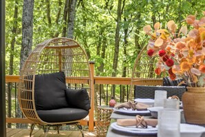 A wicker chair with a black cushion, a wicker table with a vase of flowers, and a view of a forested area. - Pocono cabin where the host thought of everything, so you don’t have to! (Bushkill)