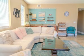 Un salon comprenant un canapé d’angle, une table basse en verre, un fauteuil turquoise et un coin cuisine avec un mur d’accent bleu.