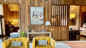 Un salon moderne avec des murs recouverts de panneaux en bois, deux fauteuils jaunes, un canapé bleu, une table basse en marbre et un tableau représentant une grange.