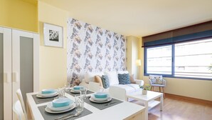 A dining area with a white table set for four, a sofa with cushions, a patterned wallpaper, and a window with blinds. - Holidays2Malaga Eslava in a closed Urbanization (Málaga)