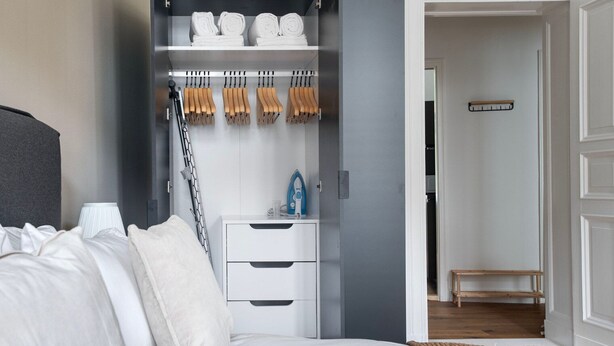 A bedroom with a bed, a bedside table with an iron, and a closet with hanging clothes and towels. - Trendy Prenzlauer Berg 1BR, nr Mauerpark, by Blueground (Berlin)