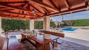 Un espacio exterior para comer con una mesa y bancos de madera, piso de piedra, y vista a una piscina y sillones de descanso.