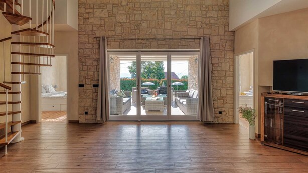 Amplia sala de estar con pared de piedra, escalera de madera, puerta corrediza de vidrio que da a un patio exterior y un televisor sobre un mueble de madera.