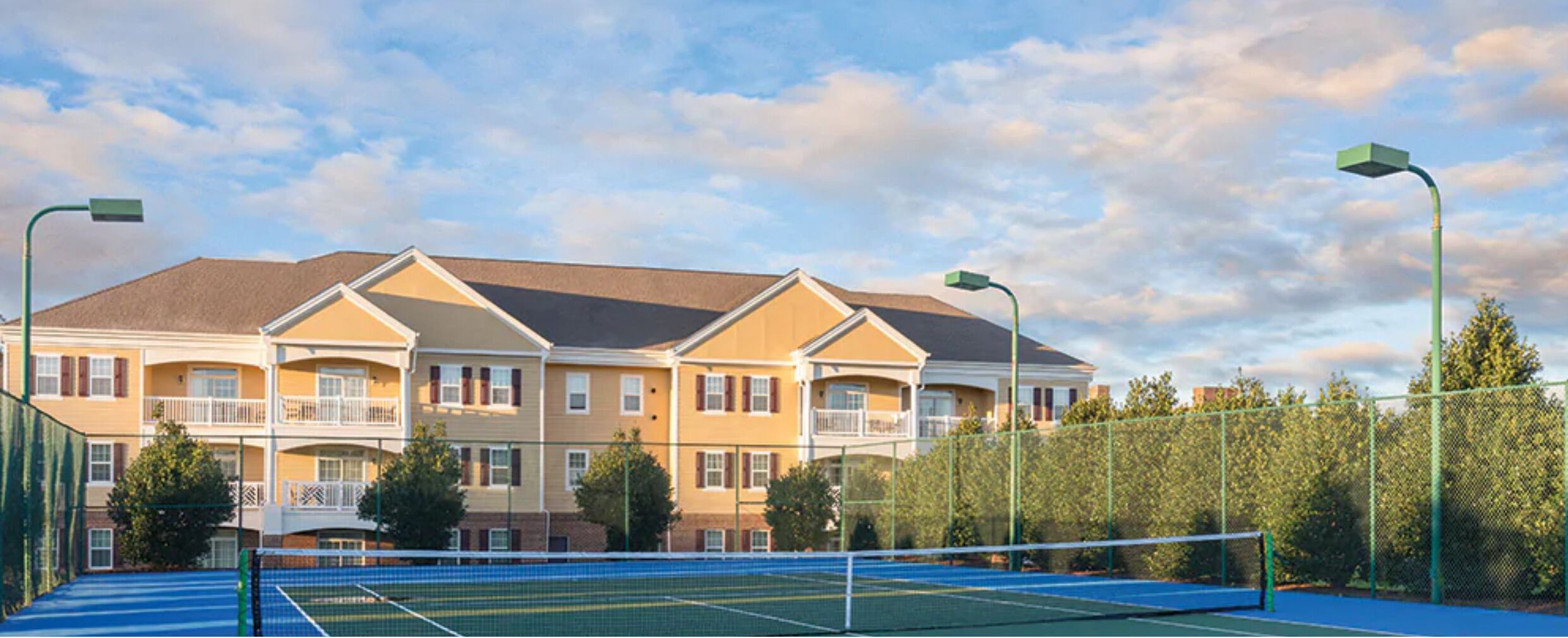 Un court de tennis avec un filet, entouré d’une clôture, et situé à côté d’un immeuble à plusieurs étages doté de balcons.
