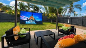 An outdoor patio with a flat-screen TV displaying a Disney castle, surrounded by black furniture and palm trees. - *Moonlight Cinema* - 15 min to Legoland! (Vista)