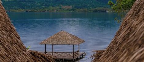 A thatched-roof hut on a dock over calm water, surrounded by lush greenery and hills.