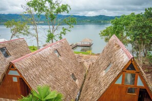 Thatched-roof cabins with wooden frames and windows, situated by a lake with a dock and a thatched-roof structure.