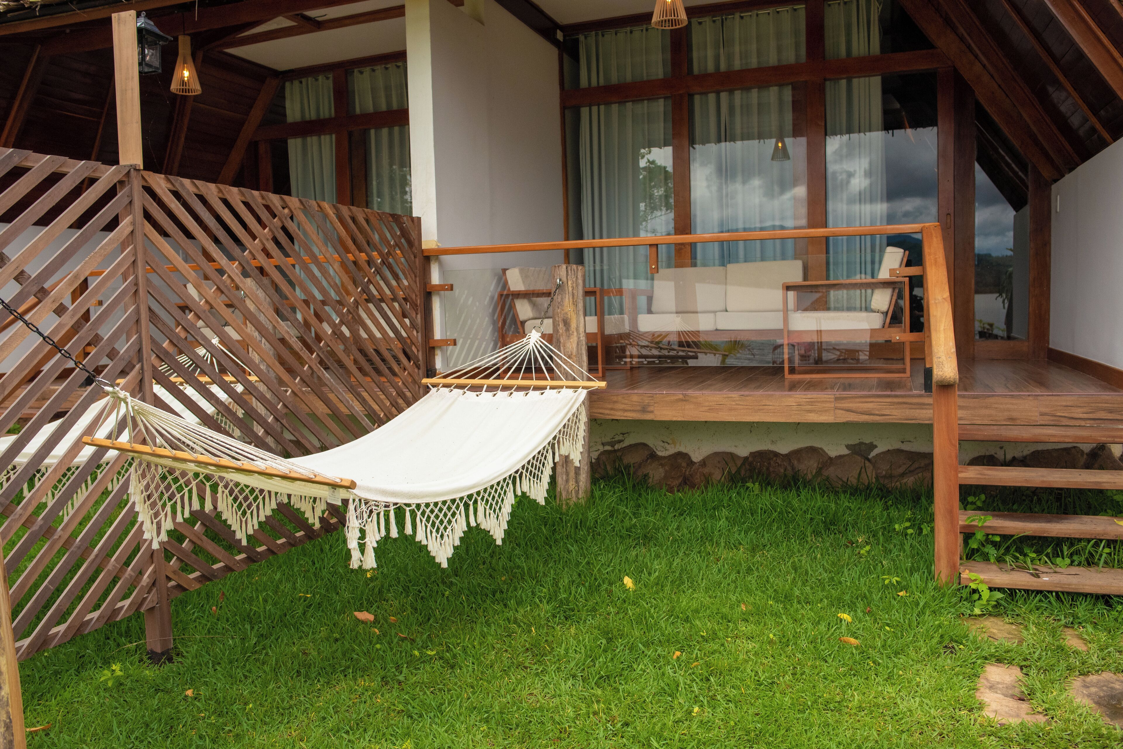 Een hangmat op een houten terras met uitzicht op een meer, een houten bankje en een glazen deur die naar een kamer leidt.