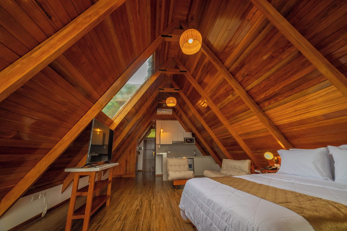Un dormitorio en el ático con vigas de madera, una cama, un escritorio y una kitchenette.
