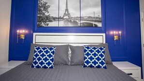 Une chambre à coucher avec un lit, deux oreillers, une table de chevet, une lampe fixée au mur et un tableau représentant la Tour Eiffel.