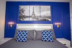 A bedroom with a bed, two pillows, a nightstand, a wall-mounted lamp, and a framed picture of the Eiffel Tower. - 4BR ChampionsGate Private Pool Lazy River Access (Davenport)