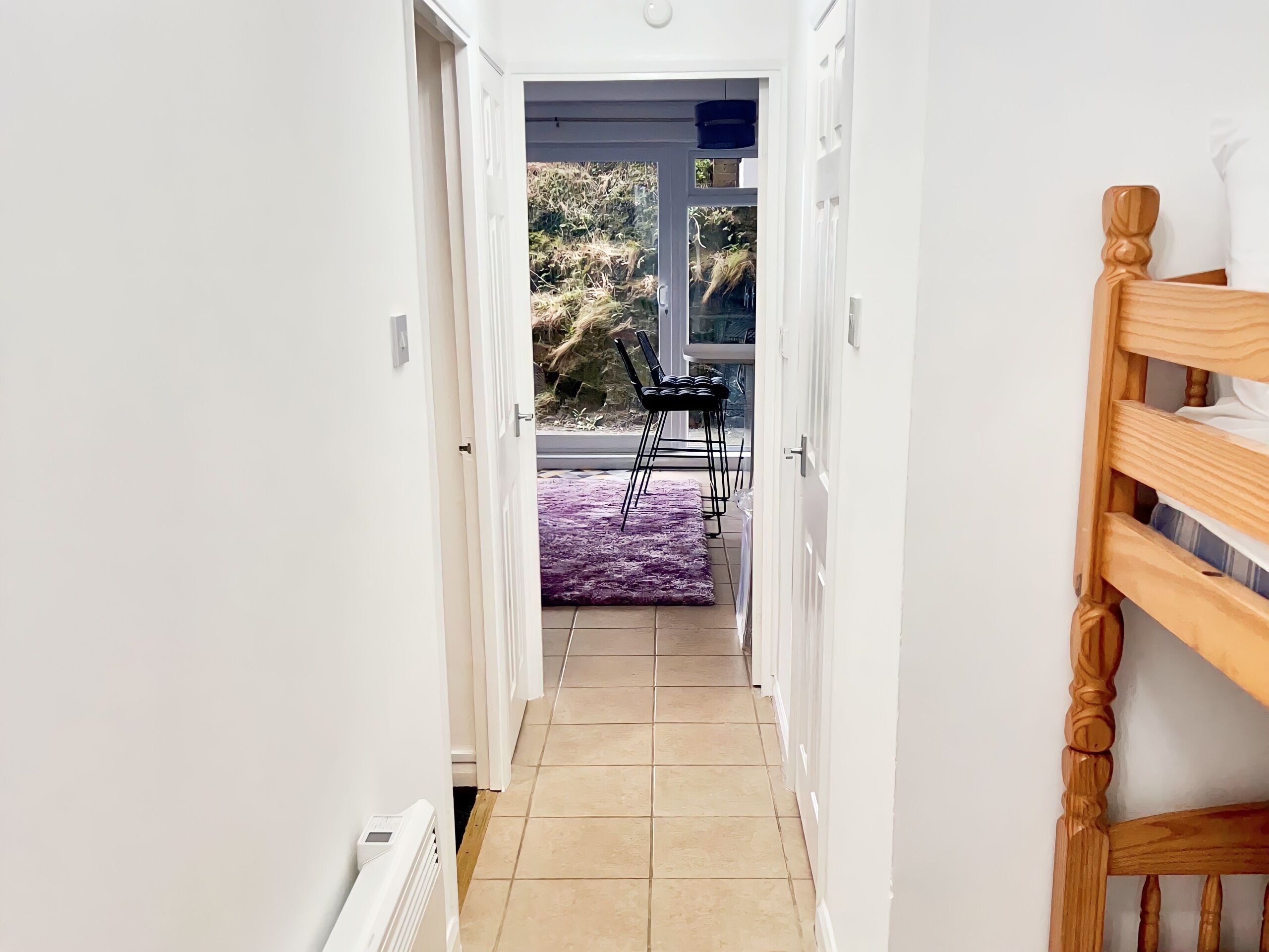 Un couloir étroit, avec un lit en bois sur le côté droit et une vue sur un patio extérieur agrémenté d’un tapis violet et de chaises.