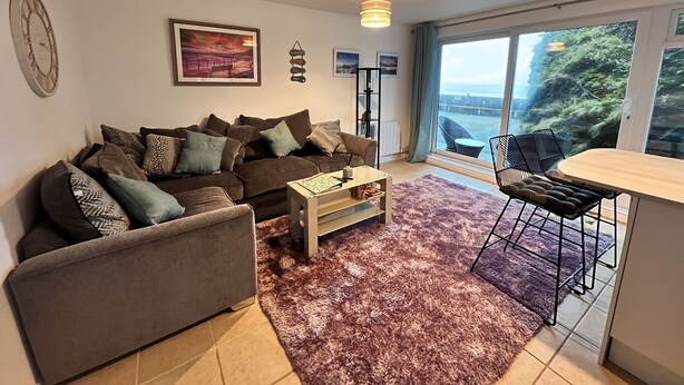 Ein modernes Wohnzimmer mit einem braunen Sofa, einem Couchtisch und einem Essbereich mit Tisch und Stühlen. Ein großes Fenster bietet Blick auf das Meer und die Stadt.