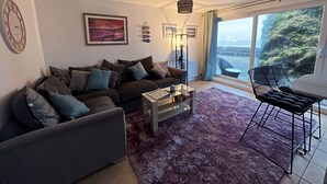 Ein Wohnzimmer mit Sofa, Couchtisch und einem Essbereich mit Stühlen. Ein großes Fenster bietet einen Blick ins Freie.