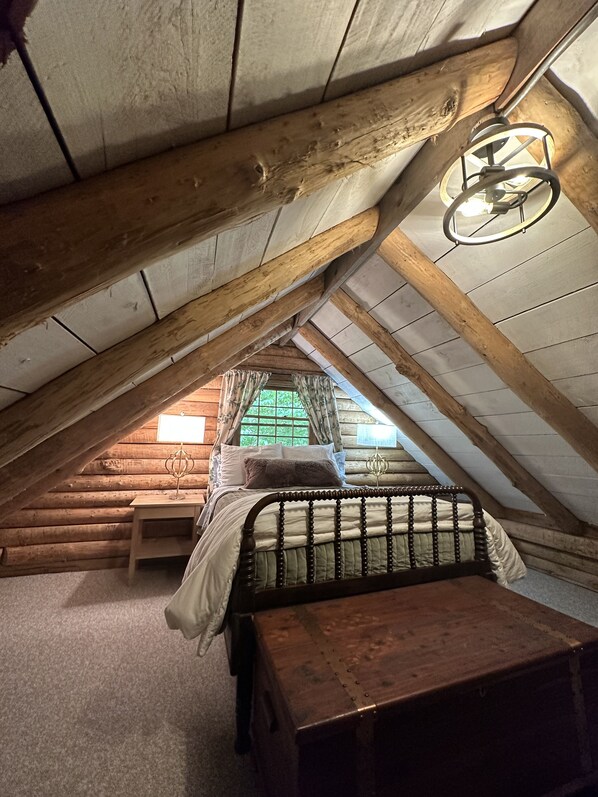Une chambre confortable située sous les toits, avec un plafond en bois, un lit doté d’une tête de lit, une petite table et une fenêtre avec des rideaux.