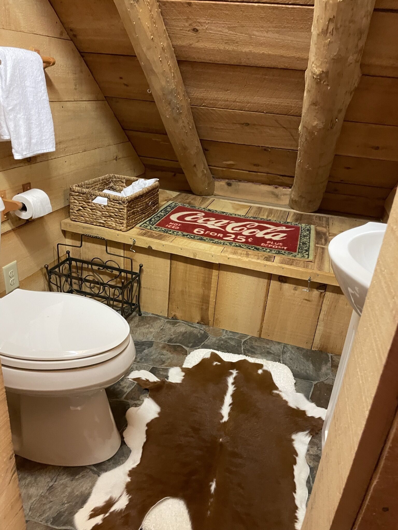 Baño de cabaña de madera con inodoro, felpudo con marca Coca-Cola, cesta tejida y alfombra de piel de vaca.