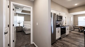 A modern kitchen with white cabinets and stainless steel appliances, a dining area with a table and chairs, and a bedroom with a bed and nightstand.