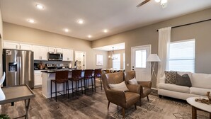 A spacious living area with a sofa, armchair, and dining table. A kitchen with modern appliances and bar stools is visible.