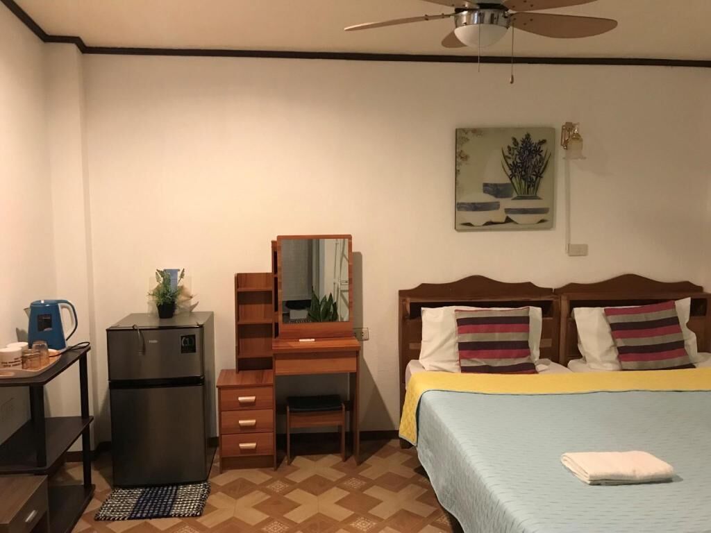 A bedroom with a bed, a desk, a refrigerator, a mirror, and a ceiling fan.