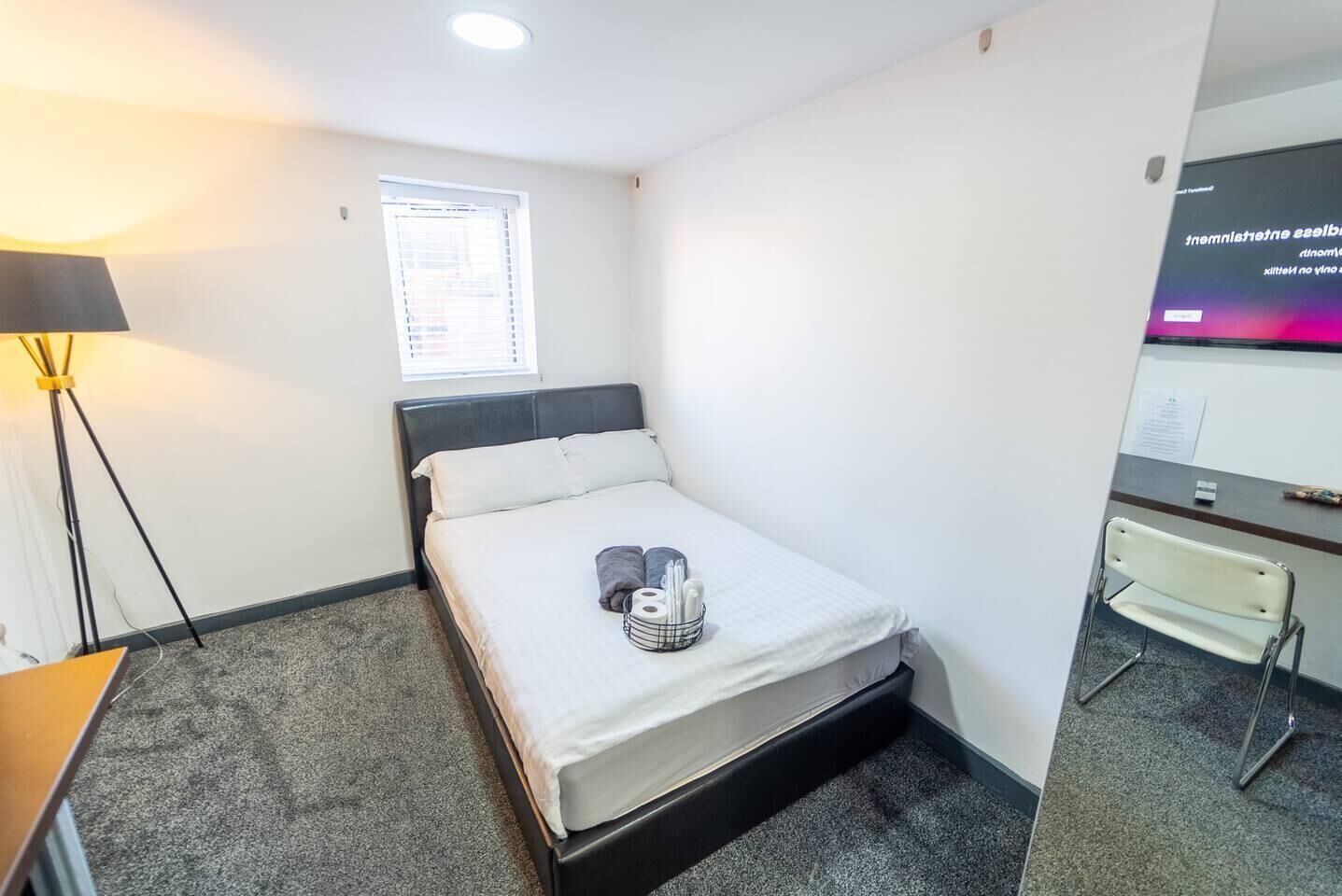 A small, neatly arranged hotel room with a single bed, a desk, a chair, a lamp, and a window with blinds.