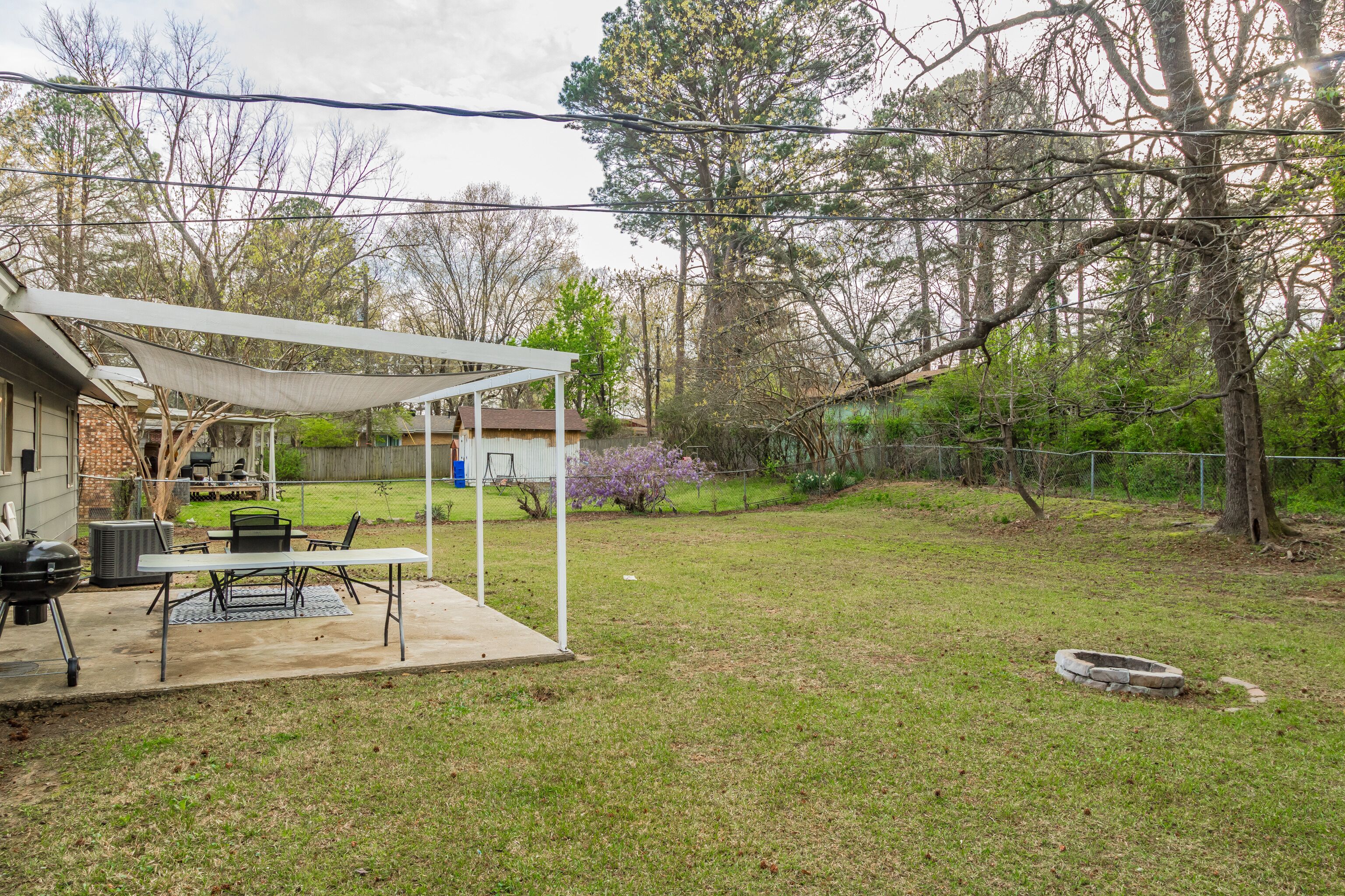 A backyard with a covered patio, outdoor grill, and a fenced area with trees and shrubs.