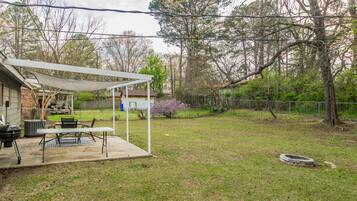 A backyard with a covered patio, outdoor grill, and a fenced area with trees and shrubs.